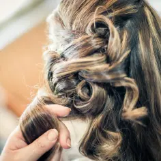 Dłoń stylisty układa loki na włosach z modnym balejażem, tworząc elegancką fryzurę.