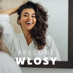 Uśmiechnięta kobieta w szlafroku podziwia swoje piękne, kręcone włosy w lustrze. Idealna pielęgnacja włosów kręconych to klucz do ich blasku.