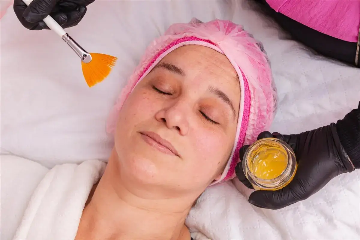 Kobieta w czepku kosmetycznym leży z zamkniętymi oczami, a kosmetyczka przygotowuje maseczkę z miodu na twarz, trzymając pędzel i słoiczek z żelem.
