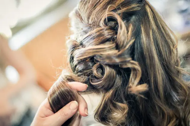 Dłoń stylisty układa loki na włosach z modnym balejażem, tworząc elegancką fryzurę.
