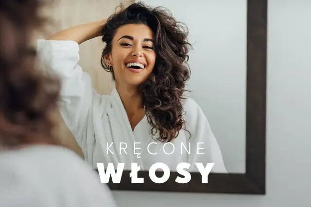 Uśmiechnięta kobieta w szlafroku podziwia swoje piękne, kręcone włosy w lustrze. Idealna pielęgnacja włosów kręconych to klucz do ich blasku.